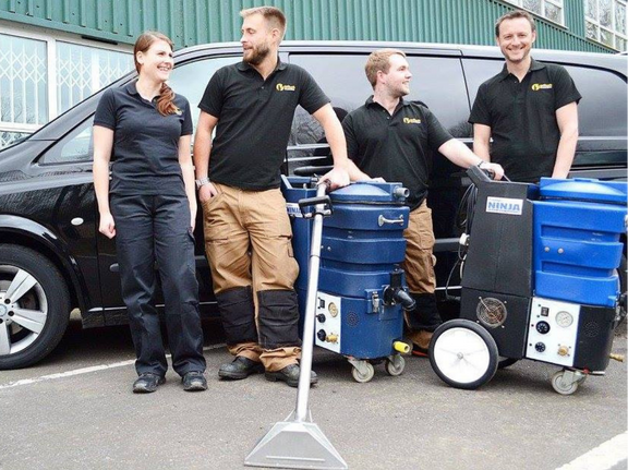 This is a photo of Chessington Carpet Cleaning carpet cleaners (three men and one woman) standing in fromt of their black van, with two steam cleaning carpet machines next to them